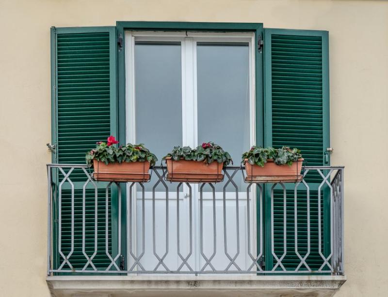 Petit balcon avec trois jardinières de fleurs accrochées à la rambarde, entre deux volets verts
